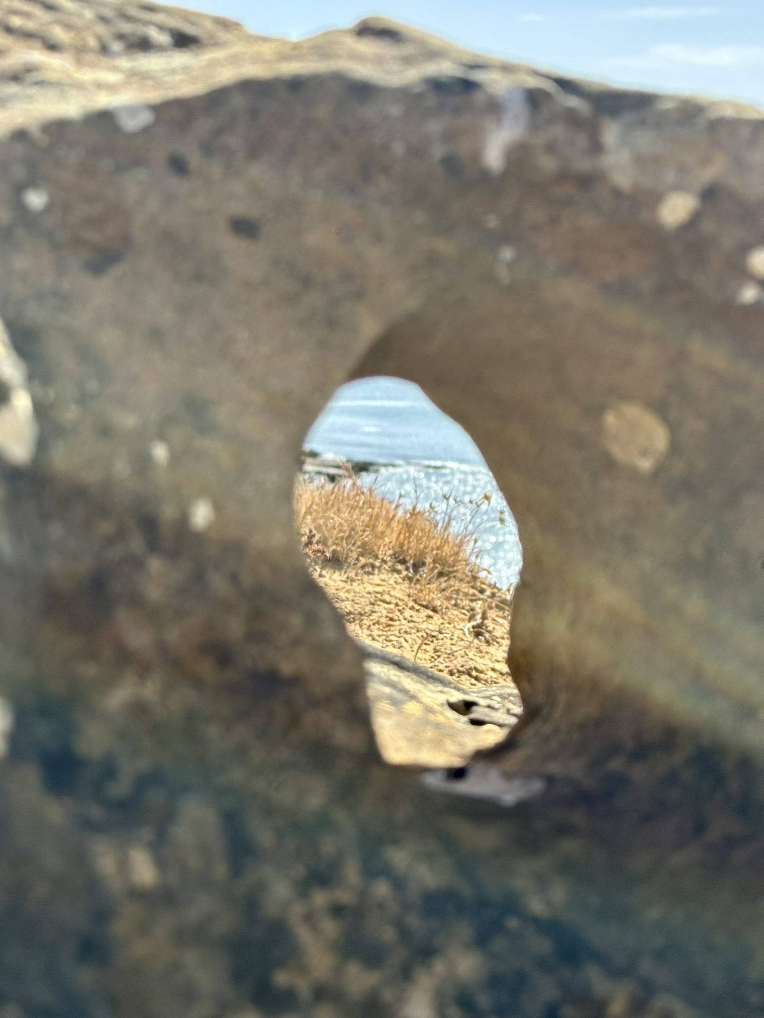 Blick durch eine natürliche Öffnung in einem großen Stein — dahinter Meer und trockenes Gras
