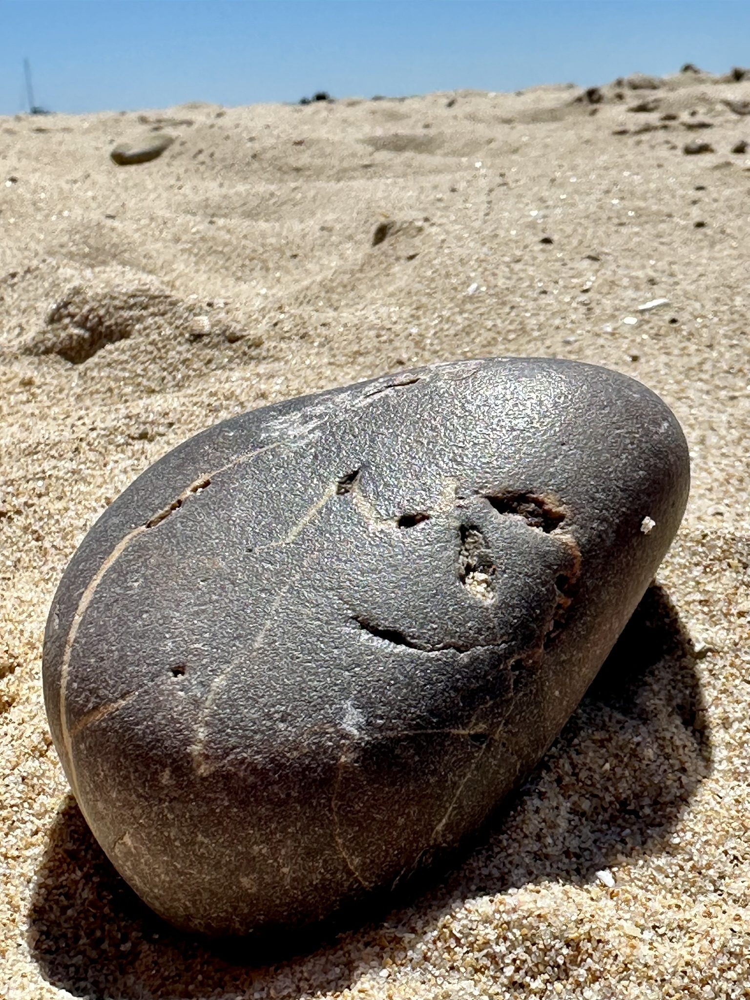 Runder dunkler Stein im hellen Sand — Vertiefungen formen Augen und ein Lächeln